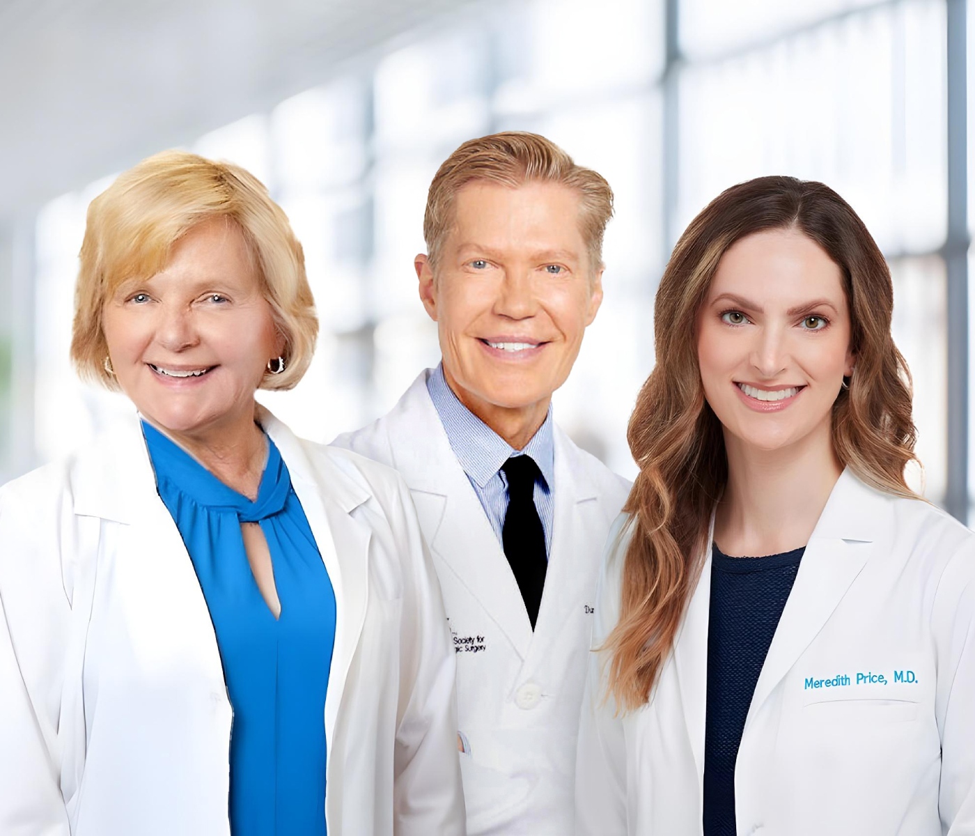 Group portrait of three smiling dermatology professionals wearing white lab coats in a modern medical office setting, with a bright, blurred background.