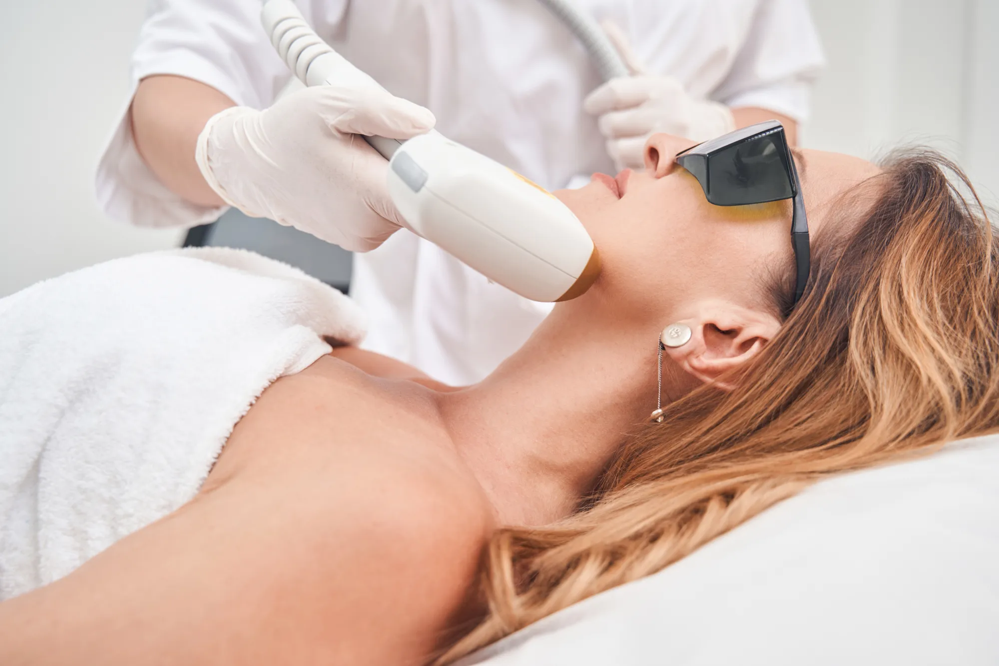 Woman receiving laser skin treatment on chin area at dermatology clinic, wearing protective goggles.