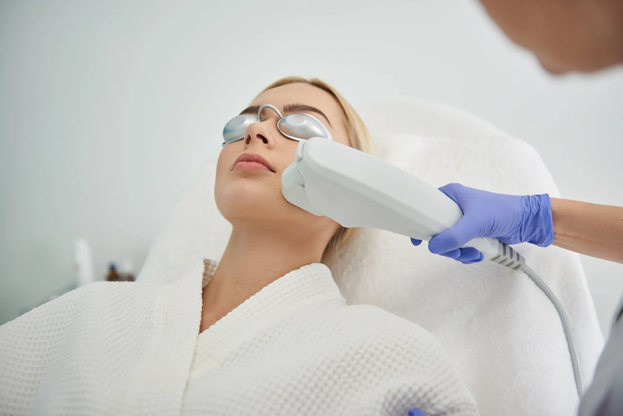 Blonde woman receiving laser facial treatment with protective goggles at aesthetic clinic.